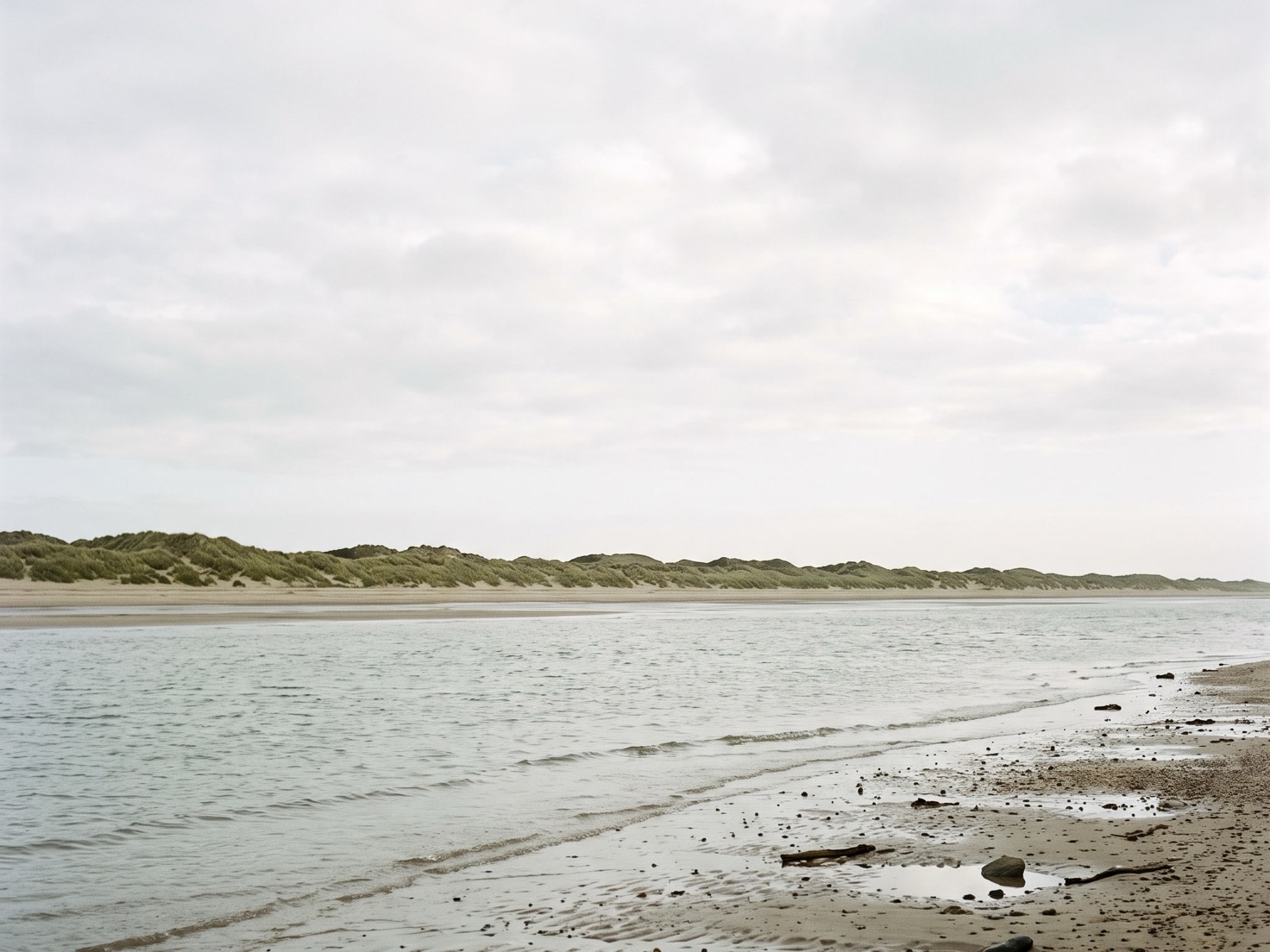 Een geul met kalm grijs water tussen nat strand en een lange rij duinen met helmgras, onder een egaal bewolkte lucht.