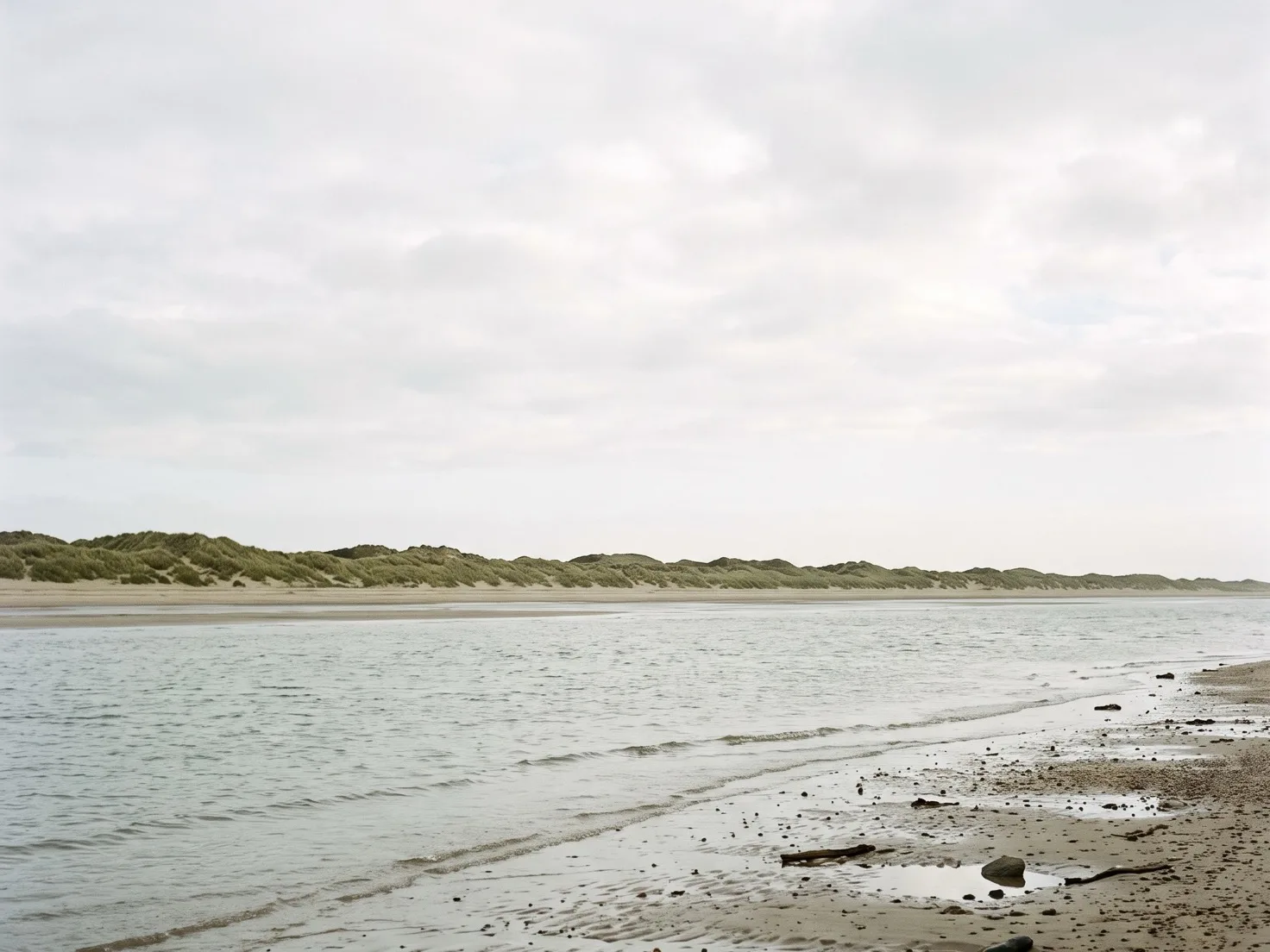 Een geul met kalm grijs water tussen nat strand en een lange rij duinen met helmgras, onder een egaal bewolkte lucht.