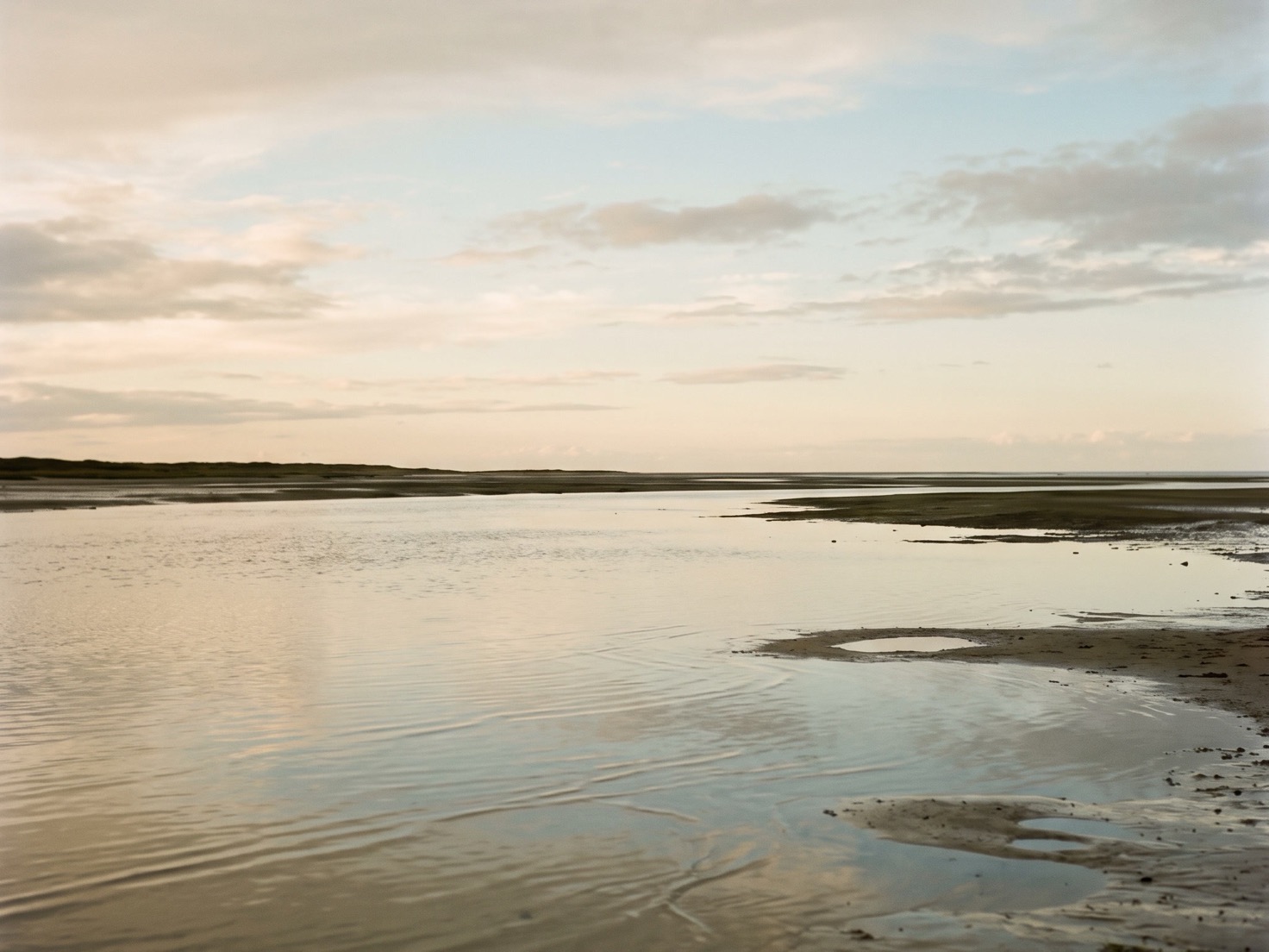 Stil ondiep water bij laagwater met zandplaten, een lage duinrand aan de horizon en een lichte lucht met zachte wolken
