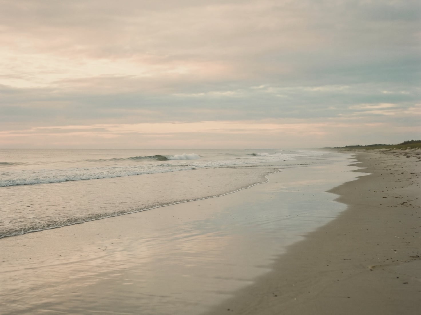 Een rustige zee bij zonsopkomst, met een vlakke golf die over glad nat zand uitloopt onder een zachtroze en grijze lucht, en rechts een leeg strand met begroeide duinen.