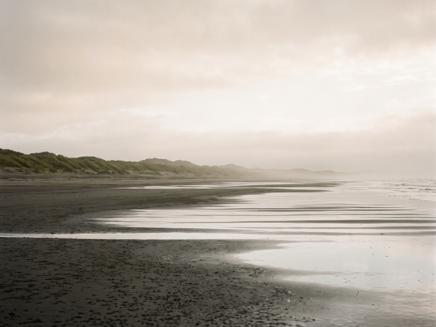 Een leeg strand bij laagwater in de ochtendmist, met ondiepe plassen die de bleke lucht weerspiegelen en een lage duinrij met helmgras in de verte.