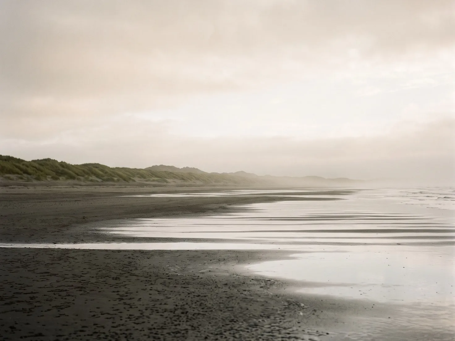 Een leeg strand bij laagwater in de ochtendmist, met ondiepe plassen die de bleke lucht weerspiegelen en een lage duinrij met helmgras in de verte.