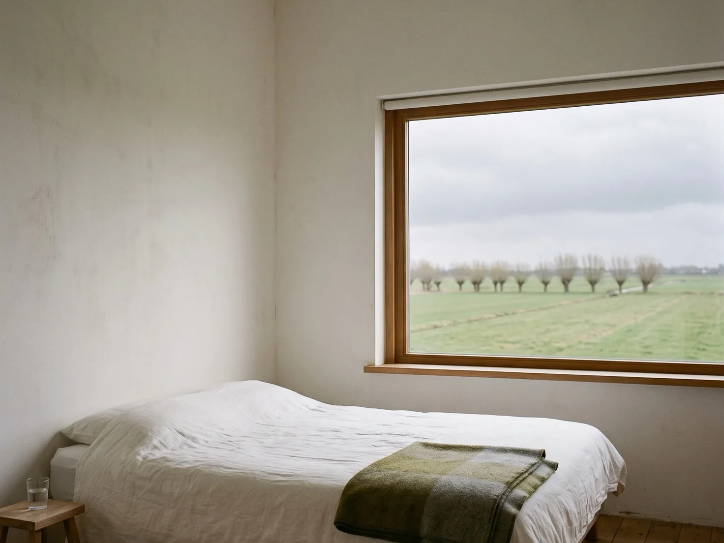 Een eenvoudige kamer met een opgemaakt bed in wit linnen en een groene wollen deken, een krukje met een glas water en een groot raam dat uitkijkt op vlakke groene weilanden met een rij knotwilgen onder een grijze lucht