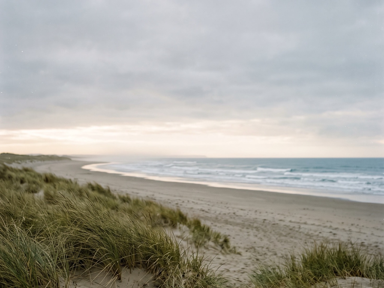 Een breed, leeg strand onder een grijze bewolkte lucht met een lichte strook aan de horizon, lage golven die aanrollen en helmgras op een duin op de voorgrond