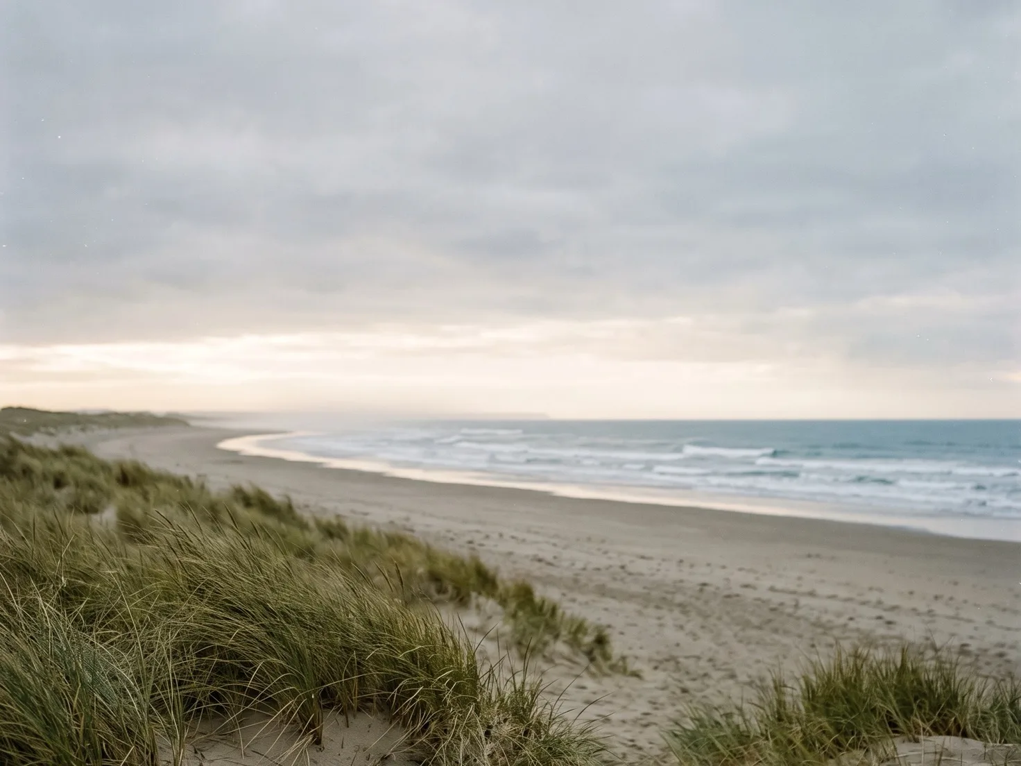 Een breed, leeg strand onder een grijze bewolkte lucht met een lichte strook aan de horizon, lage golven die aanrollen en helmgras op een duin op de voorgrond