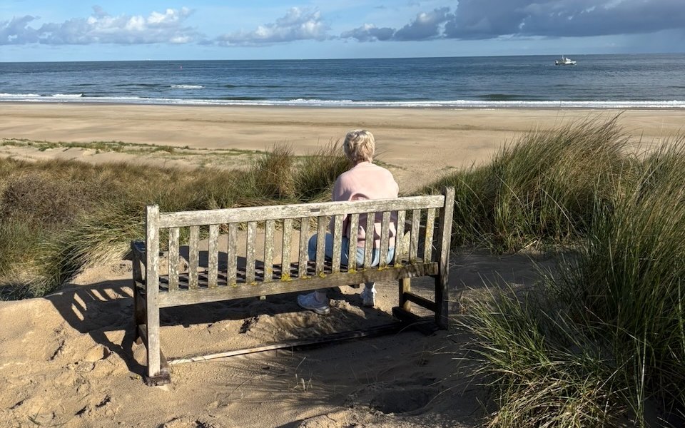 Een vrouw zit op een bankje op het duin en kijkt uit over het strand en de zee