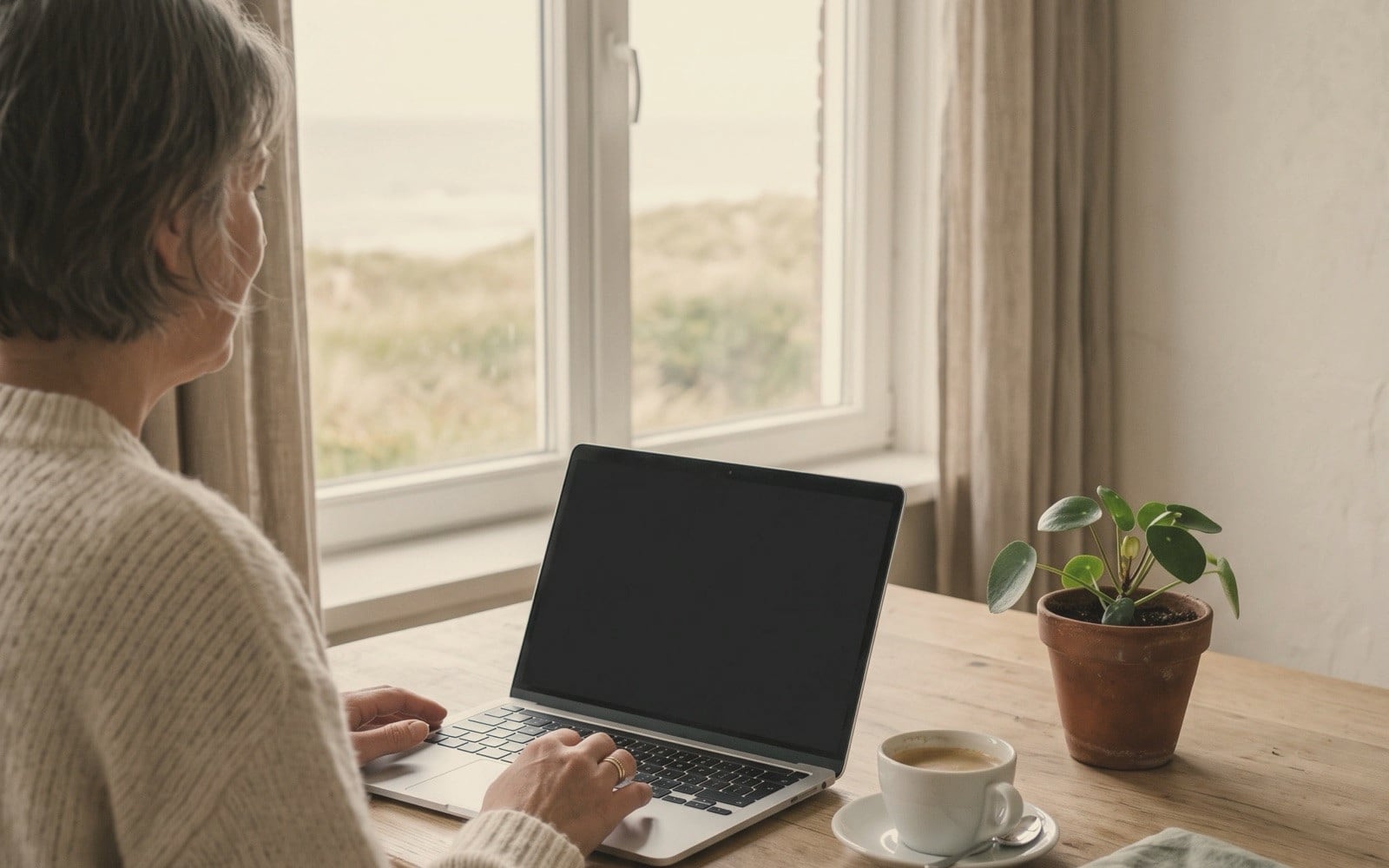 Een vrouw zit aan een houten tafel achter een laptop met een kop koffie, bij een raam met uitzicht op de duinen