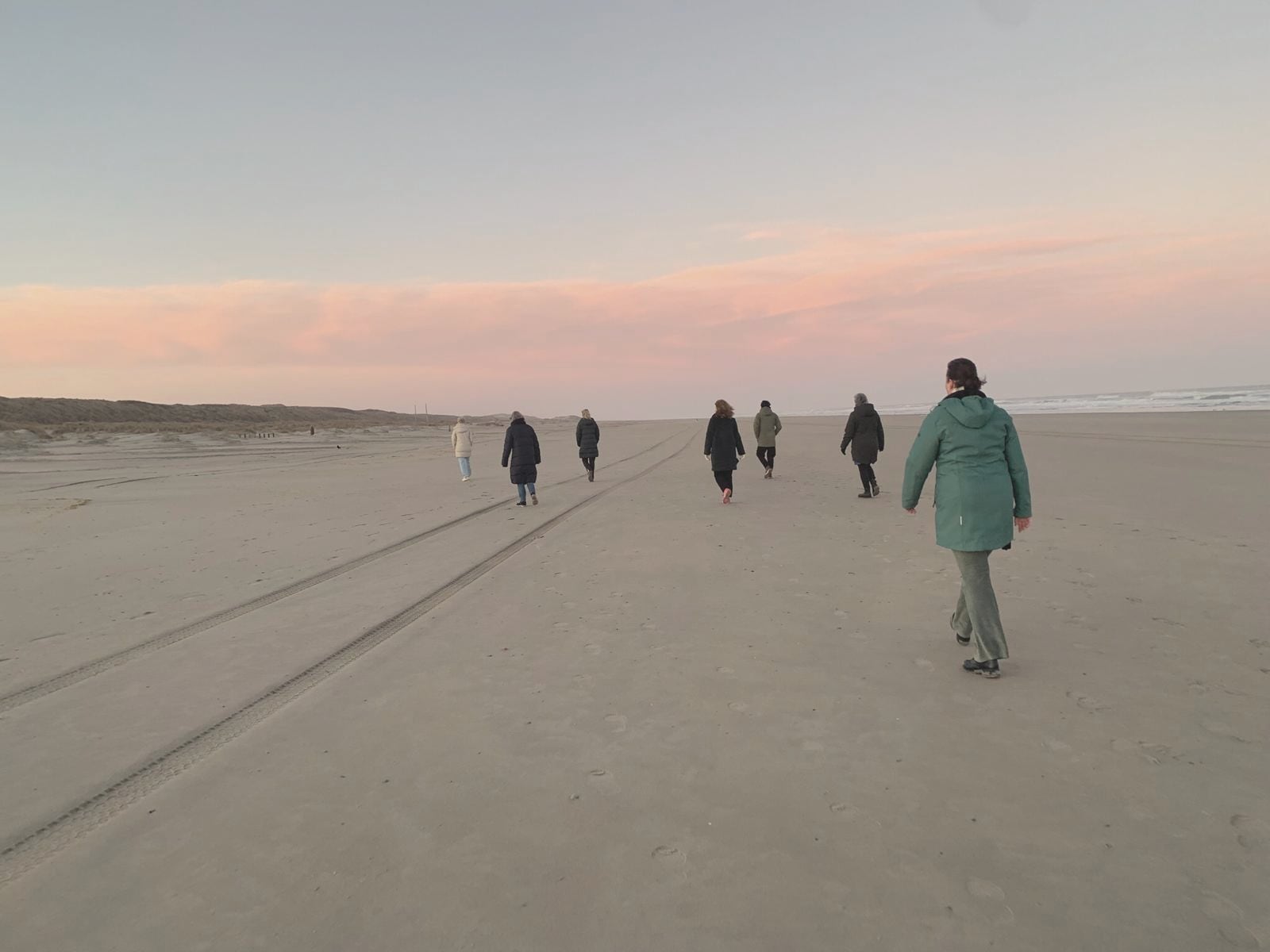 Een groep vrouwen wandelt verspreid over het brede strand van Vlieland onder een roze avondlucht